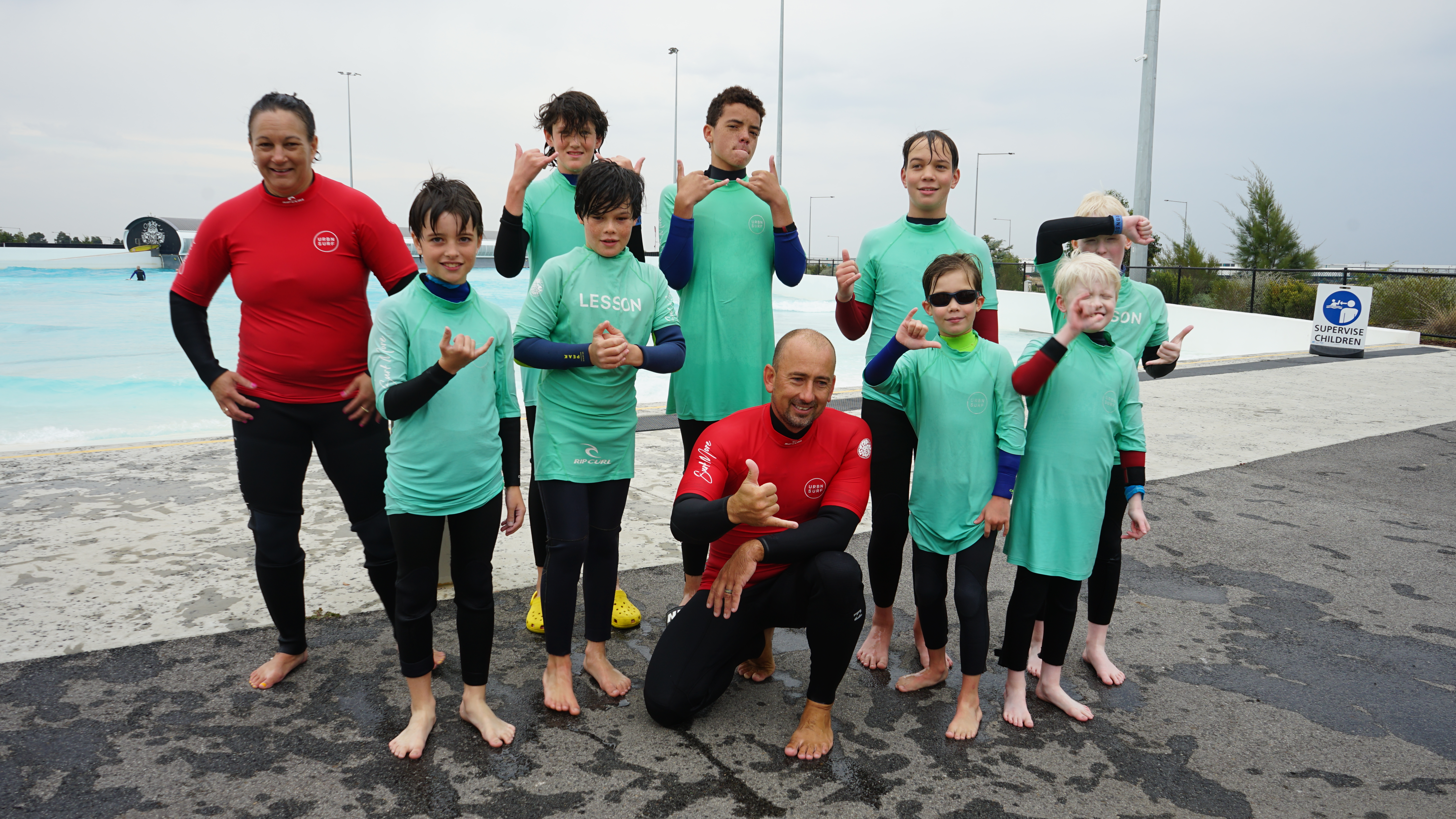 A group of children and adults in wetsuits in front of a wave pool
