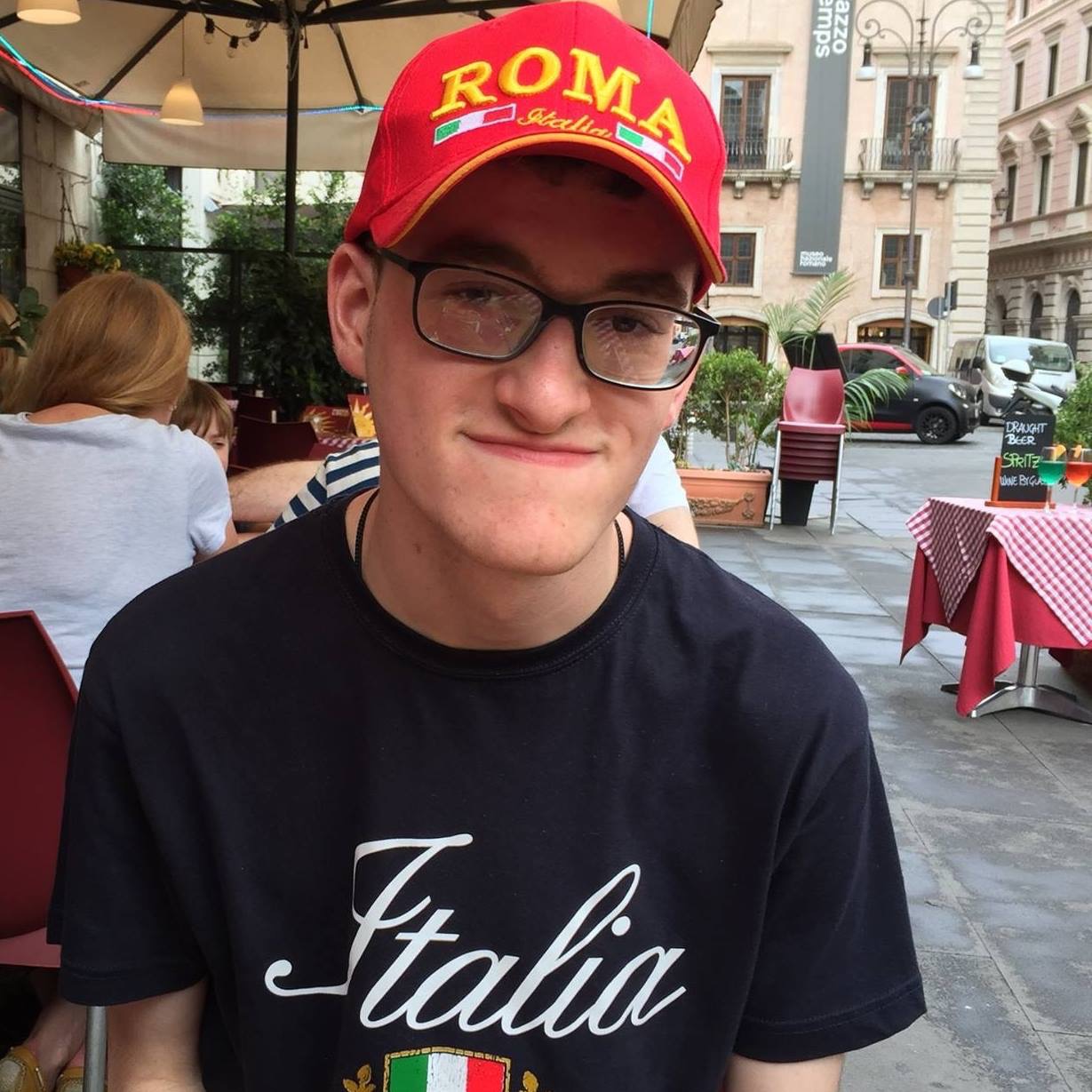 "Rohan in an outdoor cafe in Rome, wearing a red hat that reads "Roma""